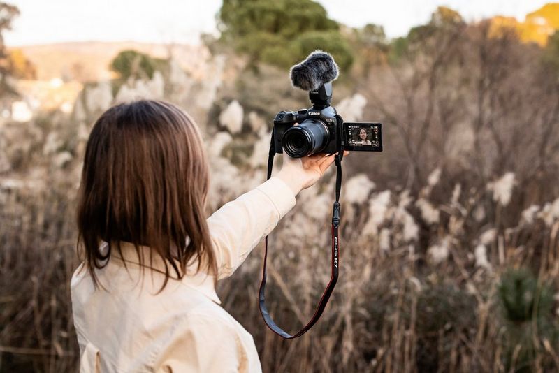 امرأة، موجهة ظهرها إلى الكاميرا، تحمل في يدها الممدودة كاميرا EOS R7 من Canon متصلة بميكروفون.