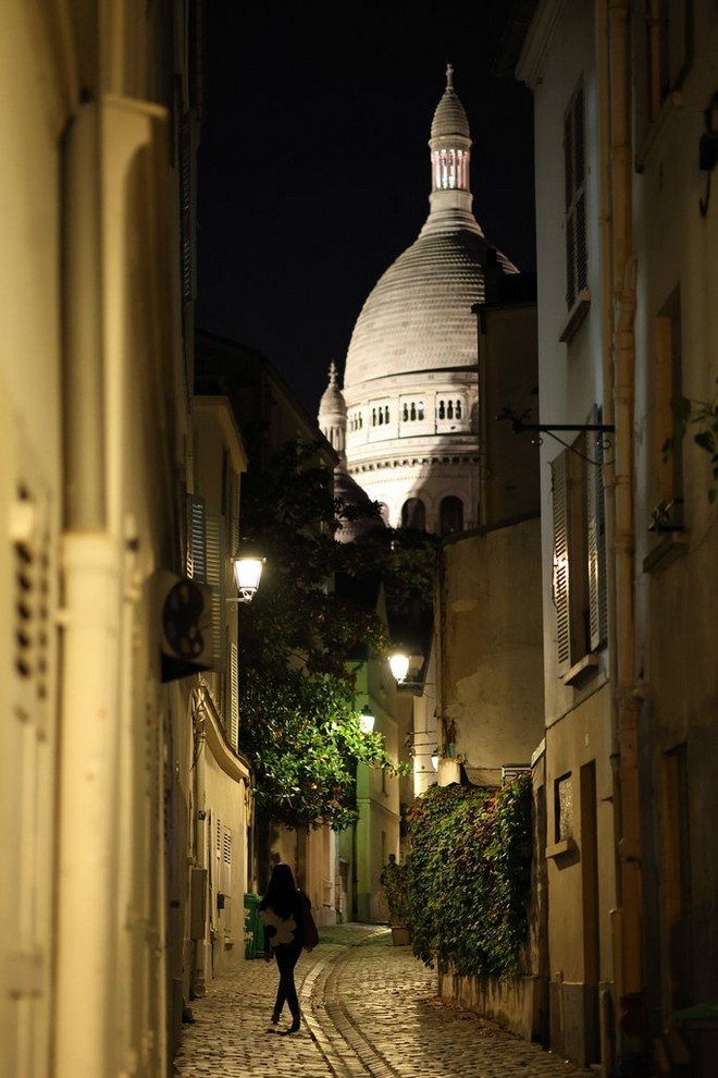 Una donna che cammina sulla strada acciottolata di Rue Saint-Rustique a Montmartre, di notte, con la Basilica del Sacré-Cœur visibile in lontananza, scatto realizzato con Canon EOS R8.