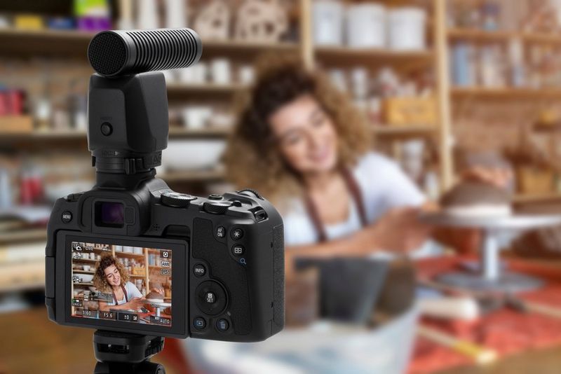 A Canon EOS R8 camera with an external microphone sits on a tripod filming a person presenting something on a raised stand.