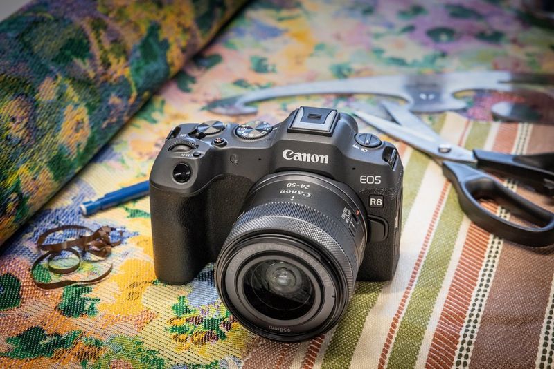 A Canon ֽ_격-8 with a Canon RF 24-50MM F4.5-6.3 IS STM lens attached, sitting on a roll of colourful fabric with a pair of scissors and a French Curve behind it.