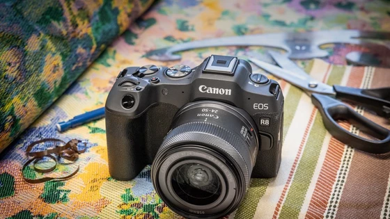 Una cámara Canon EOS R8 con un objetivo Canon RF 24-50MM F4.5-6.3 IS STM acoplado sobre un rollo de tela colorida, con unas tijeras y un curvígrafo tras él.