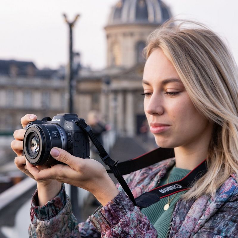 Александра Андреева держит в руках Canon EOS R8 на фоне большого размытого здания.