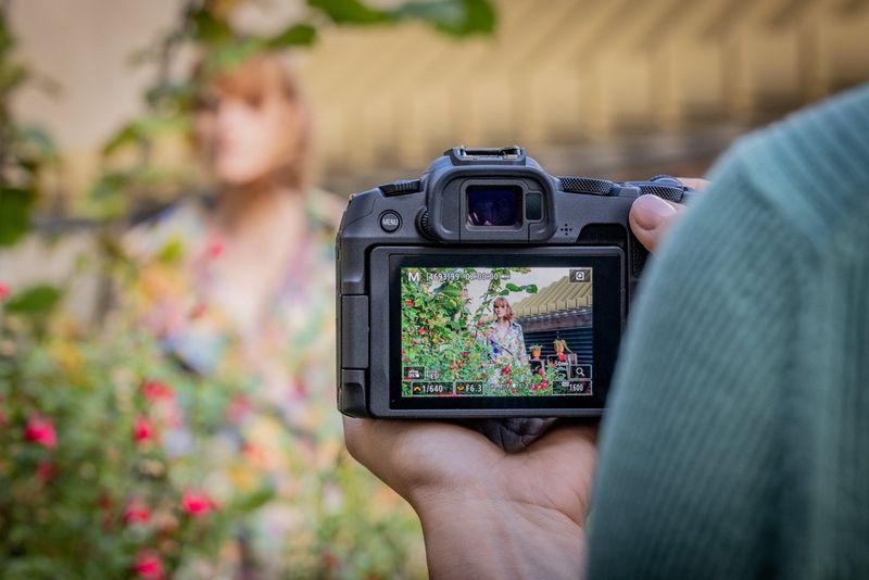 Вид заднего экрана камеры Canon EOS R8, которая используется для съемки женщины в ярком платье, стоящей рядом с цветочным кустом.