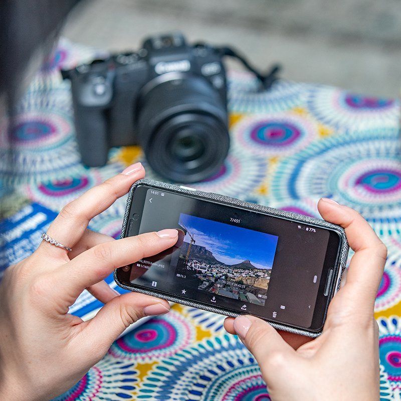 Smartphone cu o poză, ţinut în mână de o femeie aşezată la o masă cu aparatul foto în fundal.