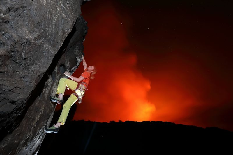 A rock climber scaling the side of an active volcano, with the red glow of the caldera behind.