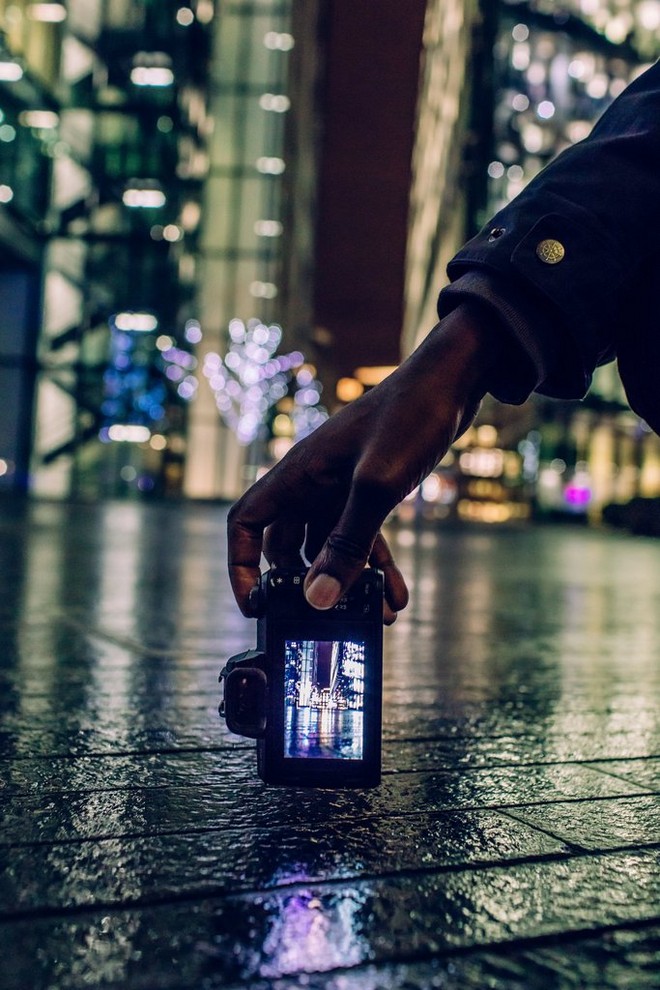 شخص يحمل كاميرا EOS M50 من Canon على ارتفاع منخفض قريب من الأرض لتصوير مدينة في أثناء الليل.