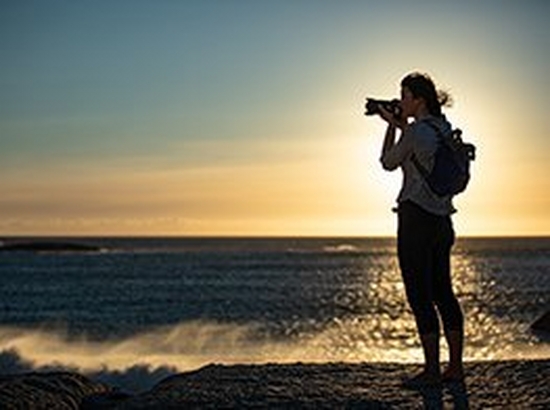 A person taking a photograph with a Canon camera and lens on a beach at sunset.