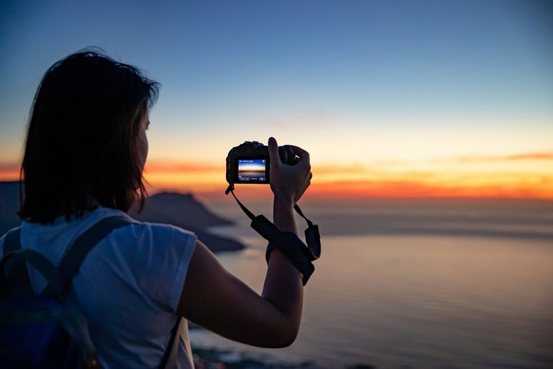 A woman in a t-shirt and backpack holds up a Canon EOS RP to take a photo of a sunrise over the ocean.