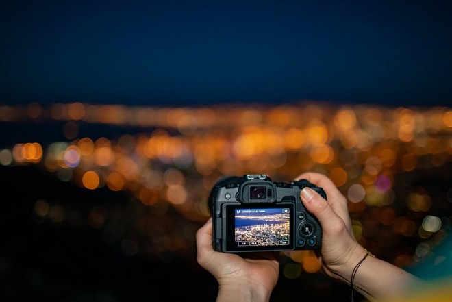 Hands holding a Canon EOS RP in front of a night-time cityscape. The scene can be seen on the camera's touchscreen.