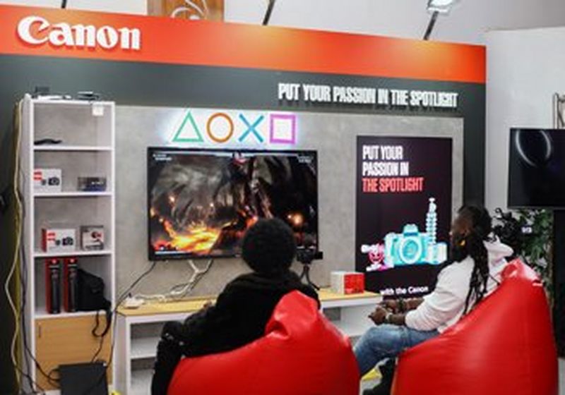 Two people sit in red beanbag gamers chairs, facing a screen mounted on a wall. One one side of the screen are shelves, holding boxes of products. On the right is a poster reading ‘put your passion in the spotlight’. Above everything is a red stripe and white Canon logo.