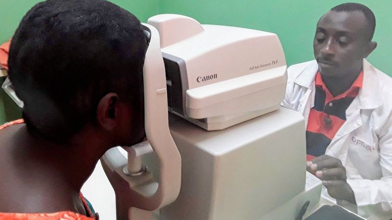 On the right, an ophthalmologist in a white lab coat and black and red striped polo shirt, looks intently at the display of a Canon Tonometer. On the other side of the machine, a patient holds their head against the machine, looking into it for their examination.