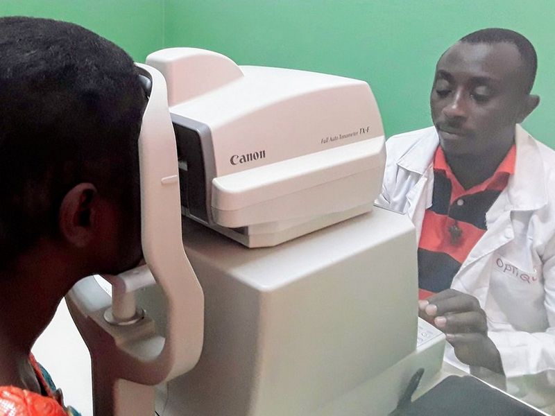 On the right, an ophthalmologist in a white lab coat and black and red striped polo shirt, looks intently at the display of a Canon Tonometer. On the other side of the machine, a patient holds their head against the machine, looking into it for their examination.