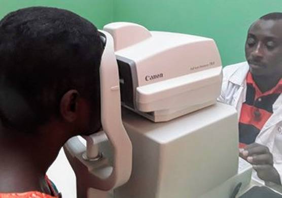 On the right, an ophthalmologist in a white lab coat and black and red striped polo shirt, looks intently at the display of a Canon Tonometer. On the other side of the machine, a patient holds their head against the machine, looking into it for their examination.