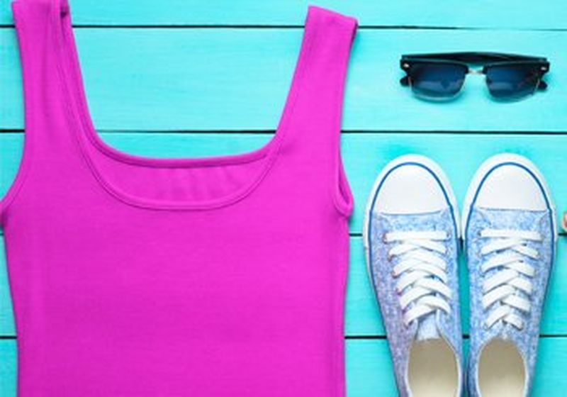 Clothing and accessories laid flat against a surface of bright blue painted wood. A violet vest, a pair of sunglasses and some blue floral converse-style plimsolls.