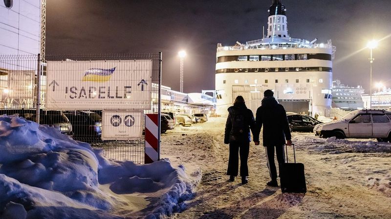 The silhouette of a couple holding hands and dragging a suitcase, as they walk through the snow towards a ferry under bright floodlights. To their left is a sign with arrows reading “ISABELLE”.