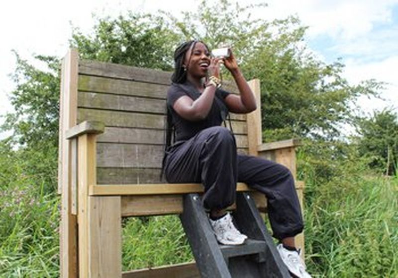 A laughing Black woman sits on a high wooden platform. She holds up a Canon PowerShot Zoom and looks through it.