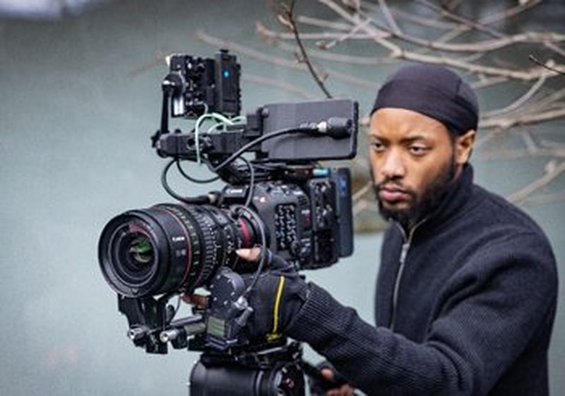 A man wearing a black zip-up fleece and black durag holds a Canon cinema camera, the bare branches of a tree behind him.