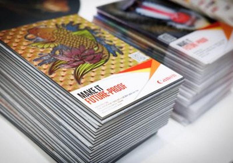 Two stacks of brochures, side by side, photographed at a slight angle from above, so you can clearly see the stack and the cover. On the cover is a Japanese-style illustration of a fish, surrounded by flowers and leaves. It is printed against a patterned background of fans. The text beneath it, in capital letters, reads, ‘make it futureproof’. Beside this text if a red, yellow and orange origami-style shape and beneath it is the red Canon logo.