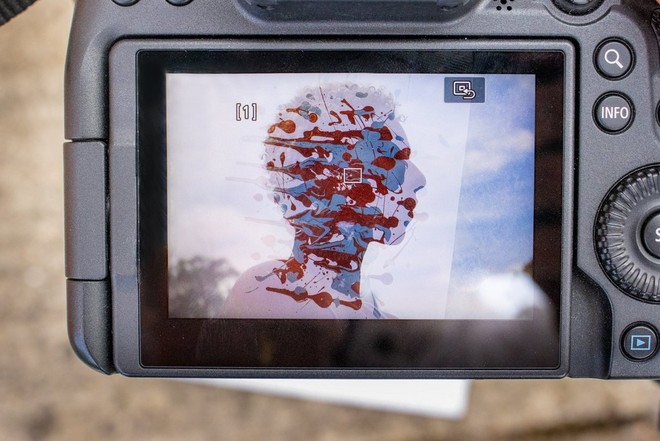 شاشة LCD على كاميرا تعرض رأس امرأة وكتفيها أمام سماء ساطعة، وتتداخل مع الصورة بقع من الطلاء الأزرق والأحمر.