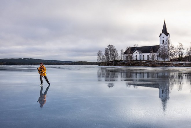 رجل يرتدي سترة صفراء يتزلج على بحيرة متجمدة. كنيسة بيضاء في الخلفية تنعكس على الجليد.