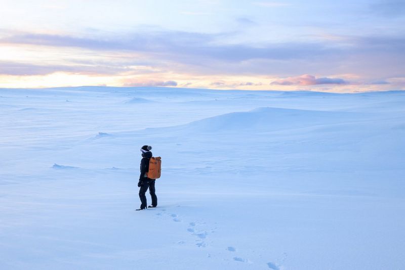 امرأة تحمل حقيبة ظهر برتقالية اللون تسير عبر منظر طبيعي مغطى بالثلوج.