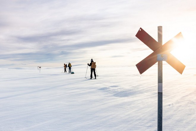 ثلاثة متزلجين عبر المناطق الريفية يجتازون مناظر طبيعية مغطاة بالثلوج. أشعة الشمس تضيء من خلفهم عمودًا يُستخدم لوضع إشارات.