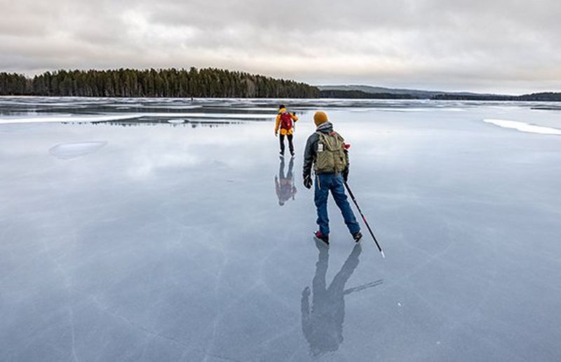 Two men carrying backpacks skate across a frozen lake.