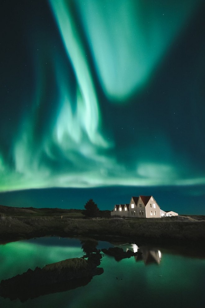أضواء خضراء رائعة للشفق القطبي فوق منزل به نوافذ مضاءة بجوار مسطح مائي كبير.