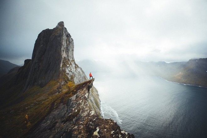 رجل يرتدي سترة برتقالية زاهية اللون ويقف على حافة منحدر يطل على وادٍ نرويجي رائع.