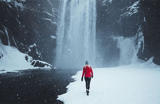 A woman dressed in a bright red winter jacket walks towards the Skógafoss waterfall in Iceland, as heavy snow falls around her.