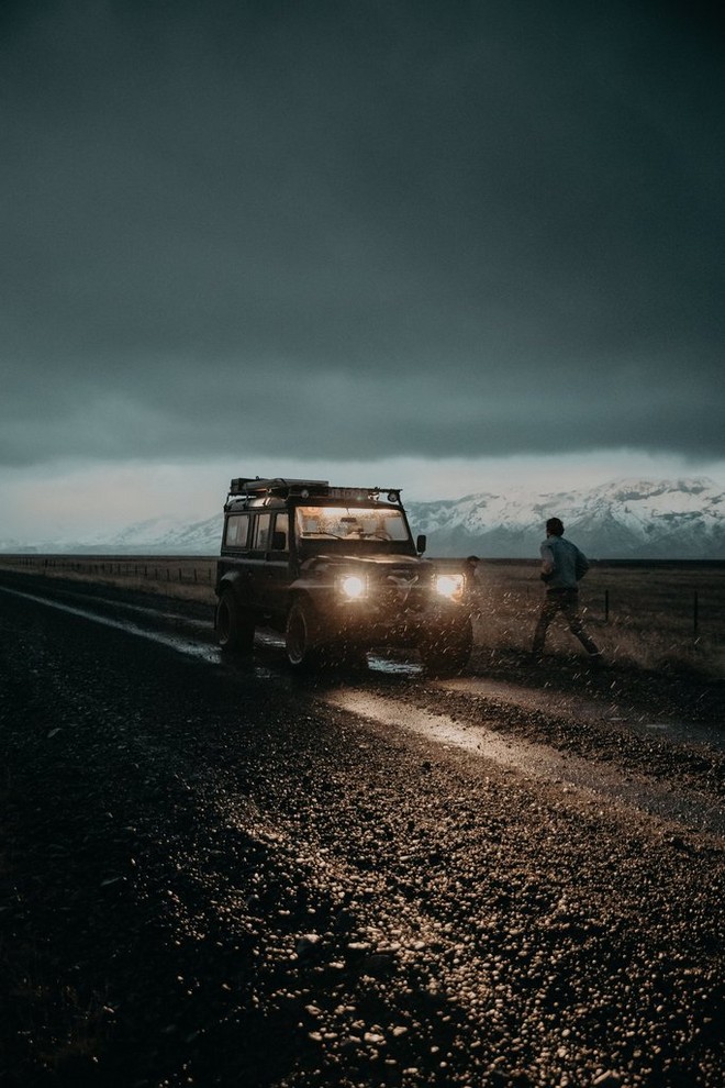 رجل يركض باتجاه سيارة جيب بانتظاره ليلاً في أيسلندا. وتضيء المصابيح الأمامية لسيارة الجيب، وتنير طريقًا مغطى بالثلوج الموحلة.
