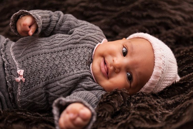 A baby wearing pink cap and grey/brown sweater lying on the side on brown blanket looking and smiling at the camera.