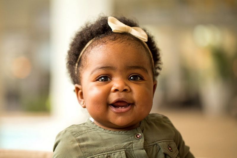 A black baby girl wearing a pink bow on her head is smiling straight to the camera. The natural light coming from a side window is spread across her face evenly.