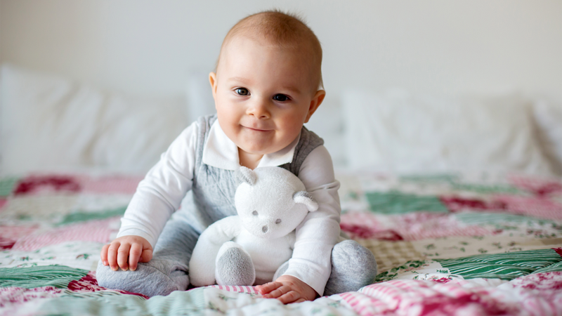 A baby holding a teddy bear sitting on a patchworked quilt on the bed, smiling straight to the camera. © Tomsickova Tatyana / Shutterstock