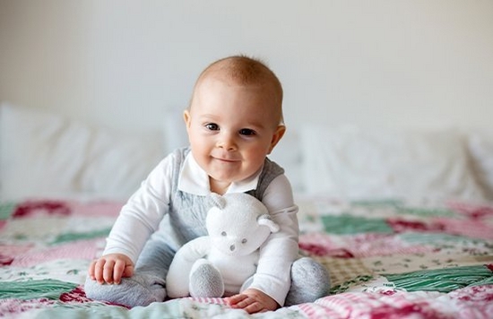 A baby holding a teddy bear sitting on a patchworked quilt on the bed, smiling straight to the camera. ? Tomsickova Tatyana / Shutterstock