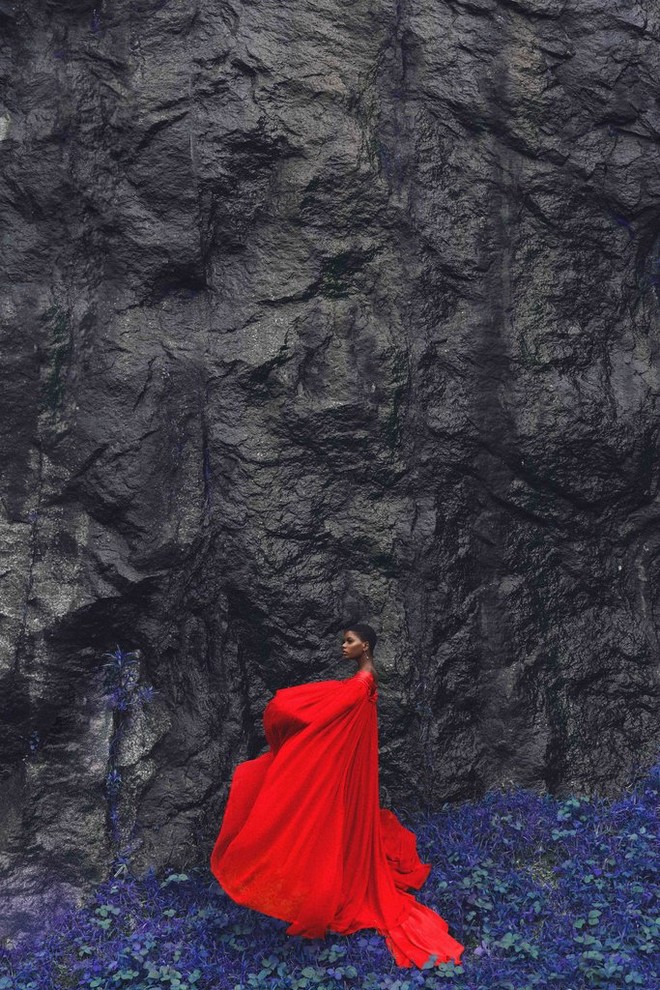 امرأة ترتدي عباءة حمراء مبهرة تقف على سجادة من الزهور الأرجوانية الزرقاء أمام صخرة رمادية داكنة.