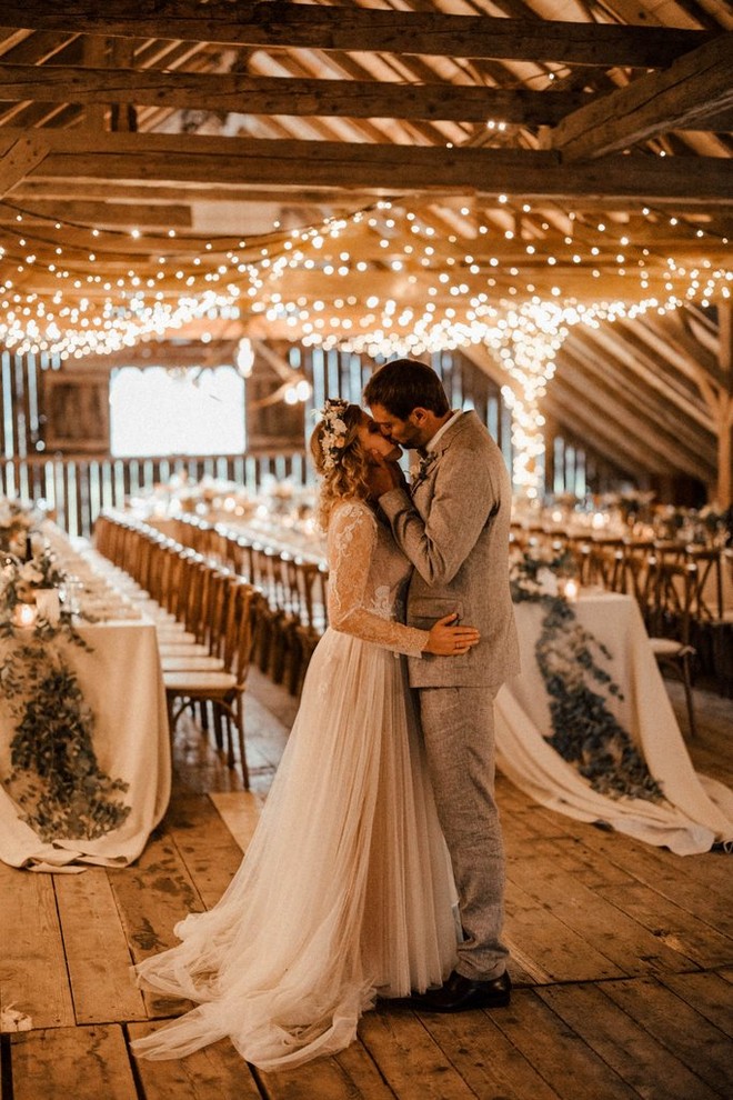 عروس وعريس يقبلان بعضهما بشغف في غرفة مليئة بأضواء الزينة والطاولات المزينة.