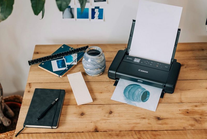 A Canon PIXMA printer prints out a picture of a small blue vase, the inspiration for which is positioned next to it.  