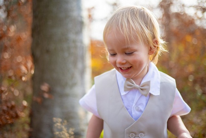 التُقطت صورة في الغابة لصبي صغير أشقر مبتسم يرتدي قميصًا أبيض قصير الأكمام وصدرية باللون البني الفاتح وربطة عنق على شكل فراشة.