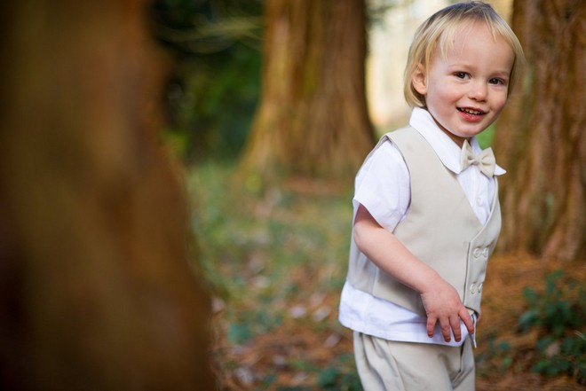 ولد صغير مبتسم يرتدي صدرية بيج وربطة عنق على شكل فراشة ويمشي بين أشجار الغابة.