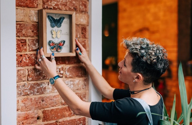 Жена закачува дрвена кутија на ѕид во која има три хартиени модели од пеперутки.