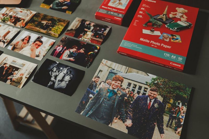 مجموعة من صور حفلة زفاف مطبوعة وحزم ورق لطابعة من Canon موضوعة على طاولة.