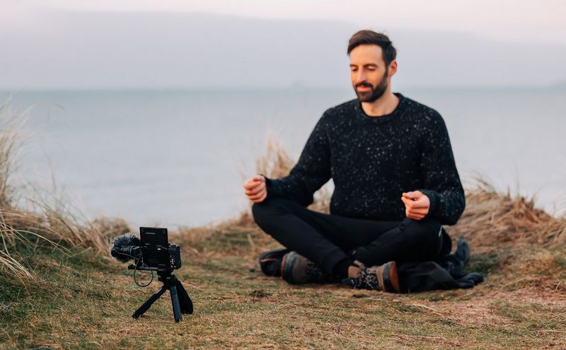 A man films himself meditating near a cliff edge with a Canon PowerShot G7 X Mark III. The ocean can be seen in the background.