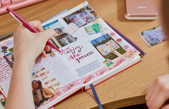 A person writing in a scrapbook, the pages filled with photos and stickers.