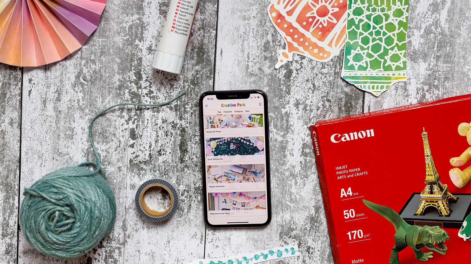 A smartphone displaying the Creative Park app surrounded by Canon printer paper, Creative Park designs and crafting tools.