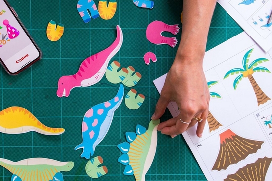 Various dinosaur templates printed from Canon Creative Park lie on a table, with a hand reaching to pick one up. 