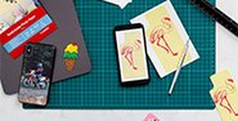 A green cutting mat with printed stickers resting on top, with a smartphone and tablet. 