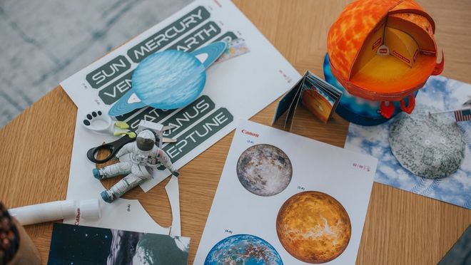 Figurica astronavta iz papirja sedi na leseni mizi, obkrožena z drugimi šablonami za izdelke iz papirja z vesoljsko tematiko iz storitve Canon Creative Park.