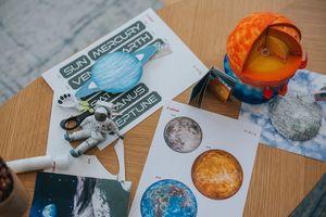 Бумажный космонавт сидит на деревянном столе в окружении других бумажных моделей на тему космоса, созданных по шаблонам из Canon Creative Park.