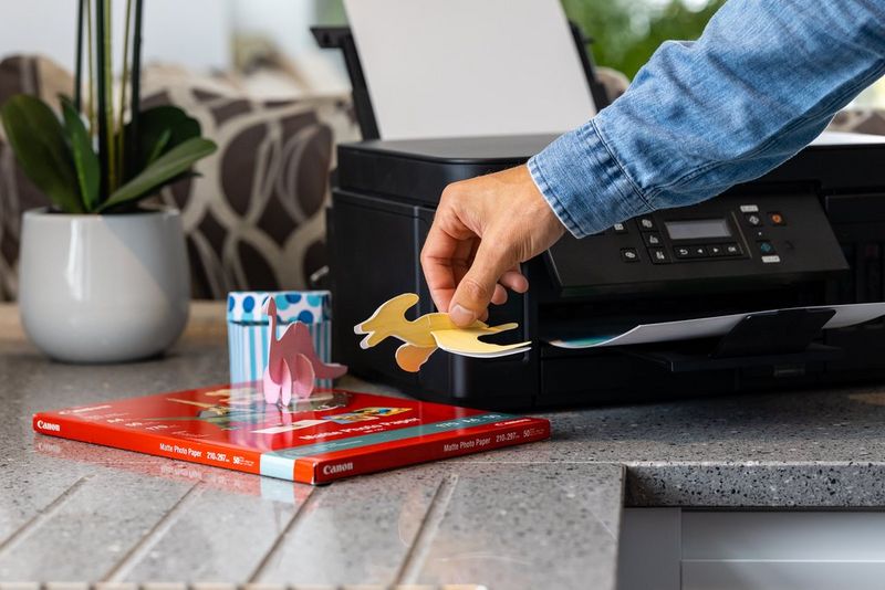 A person holding a papercraft dinosaur next to a Canon PIXMA G6040 printer.
